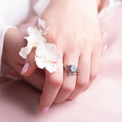 Two rings in a twig form with a Moonstone as the main stone and  White Topaz as side stones on a woman hand, which holds flowers